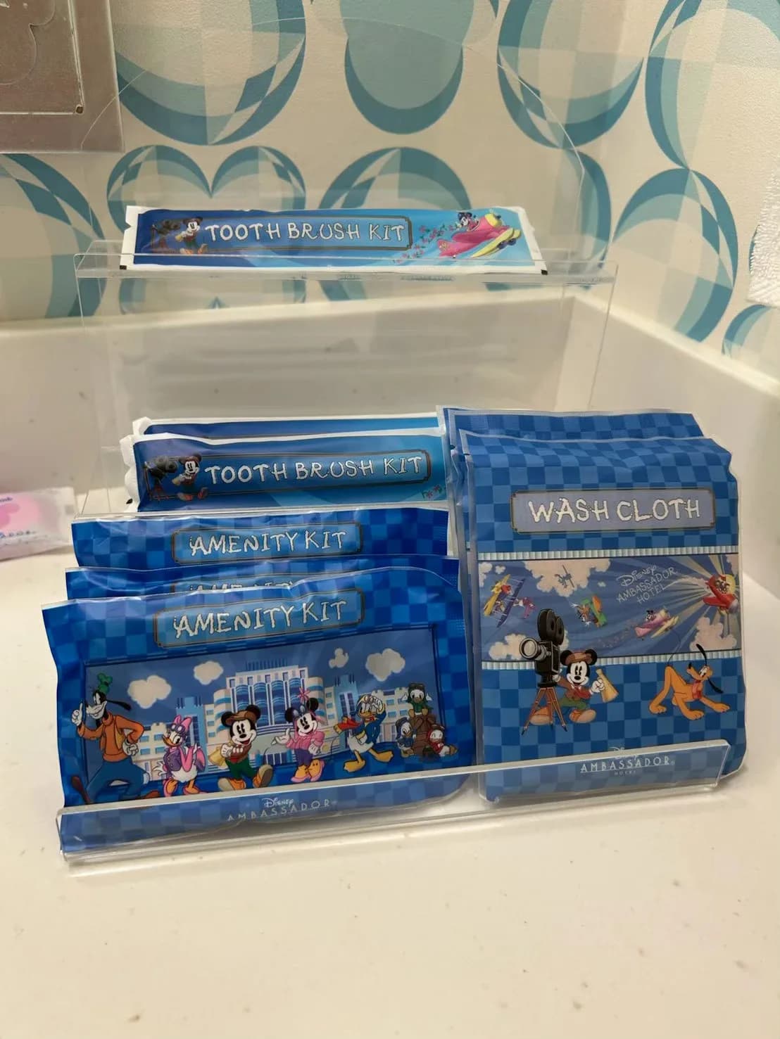 洗面台のアクリルスタンドに並ぶ TOOTH BRUSH KIT、AMENITY KIT、WASH CLOTH の袋。ミッキーたちとホテルが描かれた青いデザイン