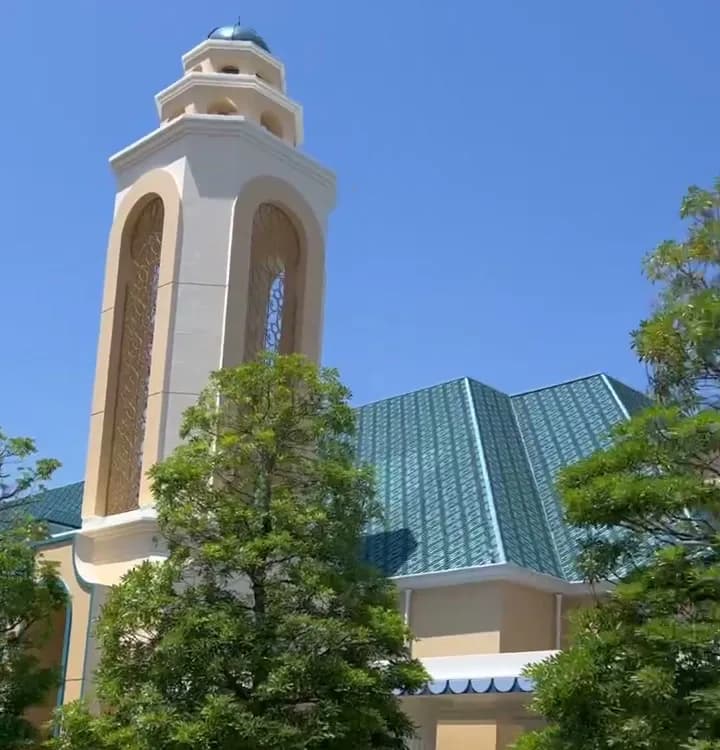晴れた日のディズニーアンバサダーホテルの外観。白い塔と青緑の屋根、手前に木々と石畳の歩道