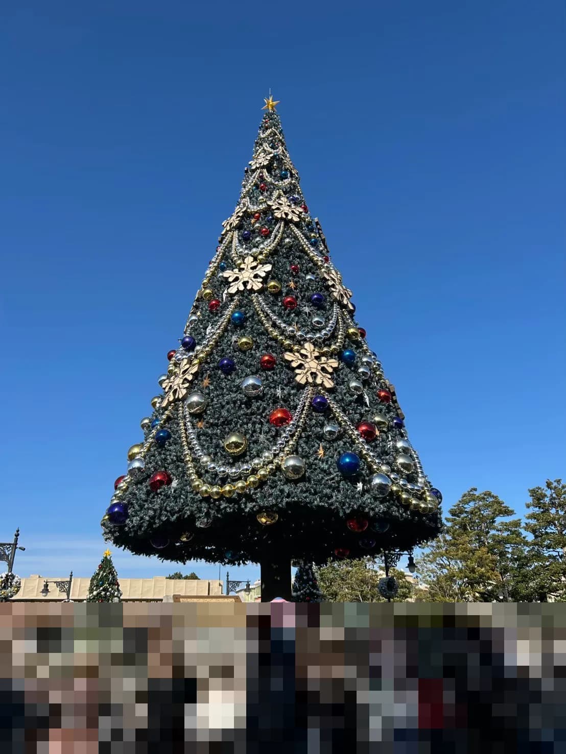 青空の下に立つ東京ディズニーシーの大きなクリスマスツリー。金色の雪の結晶の飾りと、赤・青・金・銀のオーナメントがついている。手前の人物はモザイク処理済み